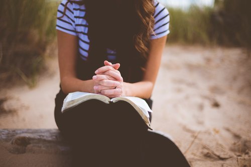 Nuori ihminen istuu hiekkadyyneillä ja rukoilee kädet avoimen raamatun päällä.