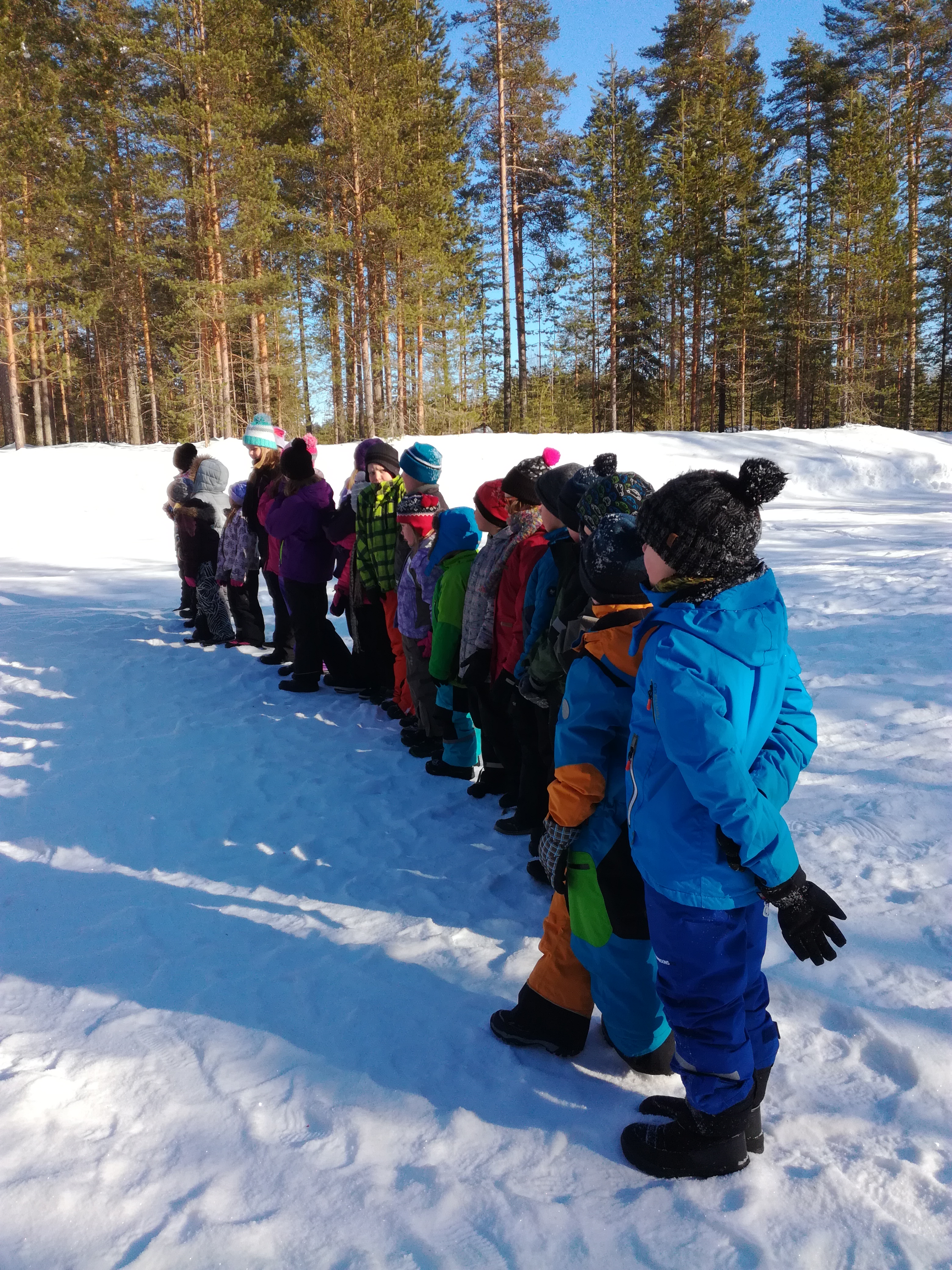 Lapset seisovat rivissä lumisella kentällä. Taustalla on kauniin sininen talvitaivas ja mäntyjä.