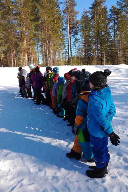 Lapset seisovat rivissä lumisella kentällä. Taustalla on kauniin sininen talvitaivas ja mäntyjä.