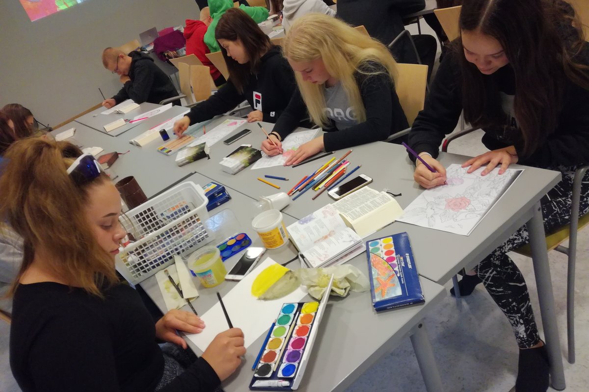 Nuoret istuvat pöydän ääressä ja maalaavat paperille taideteoksia. Osa nuorista värittää värityskuvia.