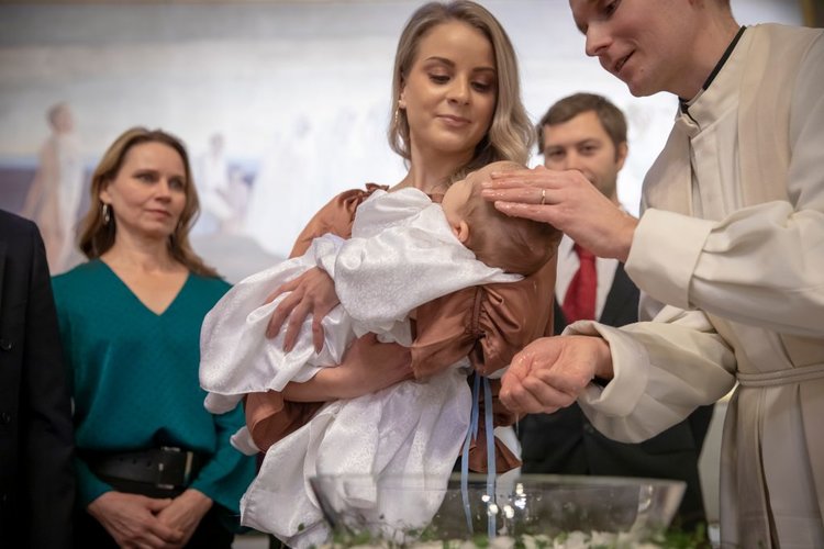 Pappi kastaa vauvaa, nainen pitelee vauvaa sylissään. Taustalla kastejuhlavieraita.
