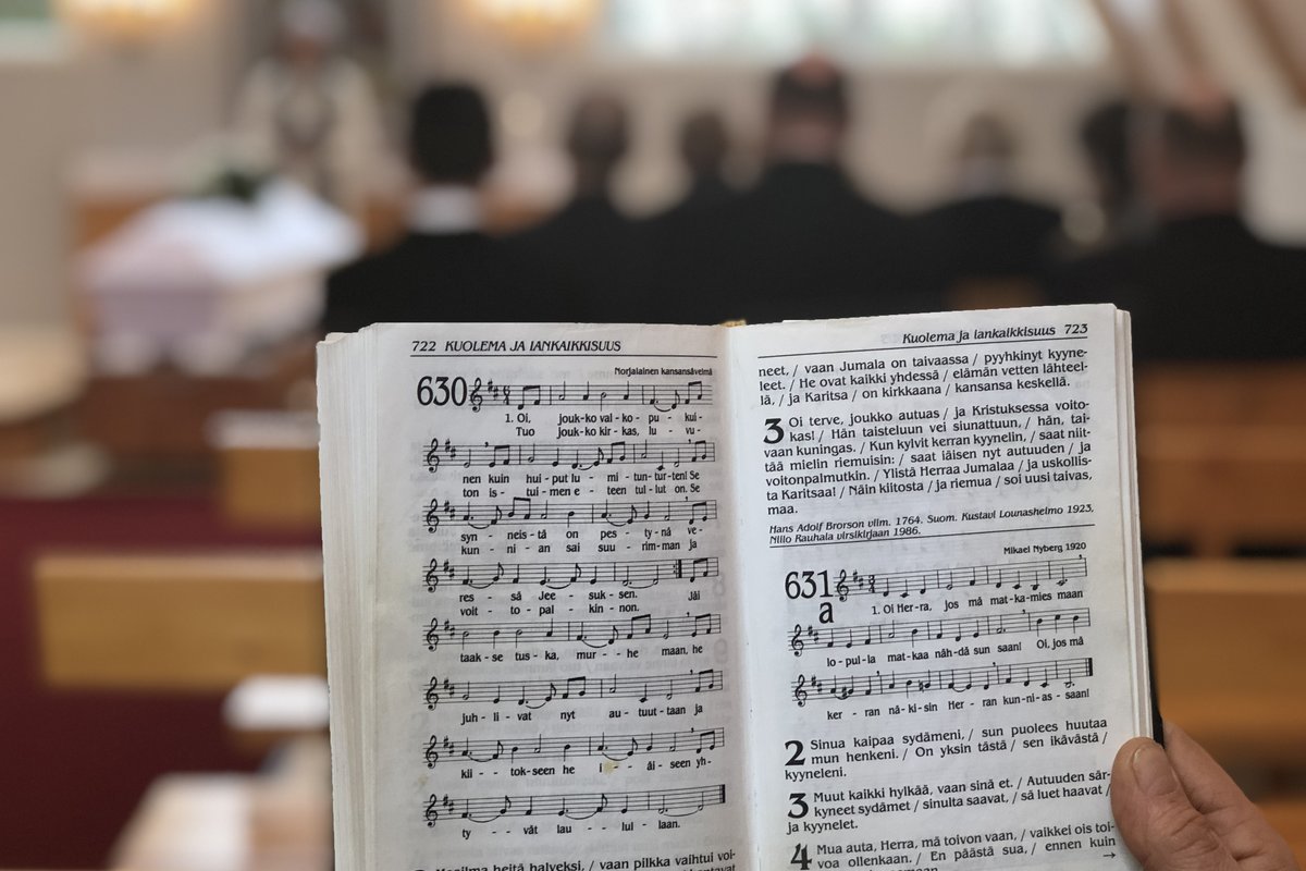 Hautajaisvieraan kädessä virsikirja, jossa näkyy hautajaisvirsi. Taustalla hämäränä siunaustoimitus.