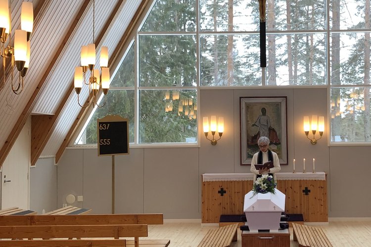 Siunauskappelissa pappi on arkun ääressä. Kappelin päädyn suuresta lasi-ikkunasta näkyy hautausmaan puita.